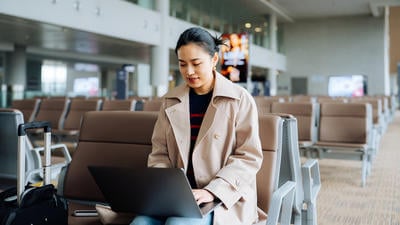 Asian businesswoman working on laptop while sitting at airport terminal waiting for her flight, a suitcase by her side