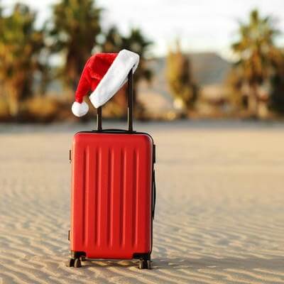 Red suitcase with Santa Claus red hat on handle with tropical sand beach, palms, mountains and sea background