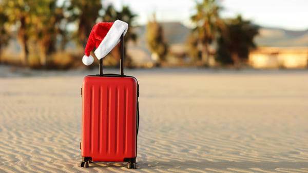 Red suitcase with Santa Claus red hat on handle with tropical sand beach, palms, mountains and sea background