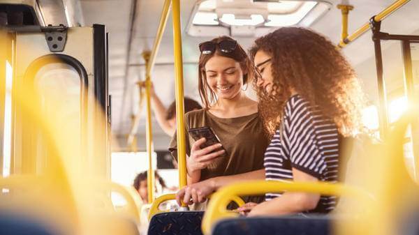 Two girls watching phone and smiling while standing on a bus
