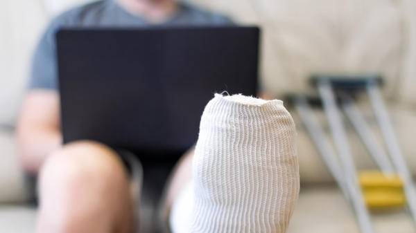A man sits on a sofa in a cast with a laptop. A leg in a cast lies on a chair